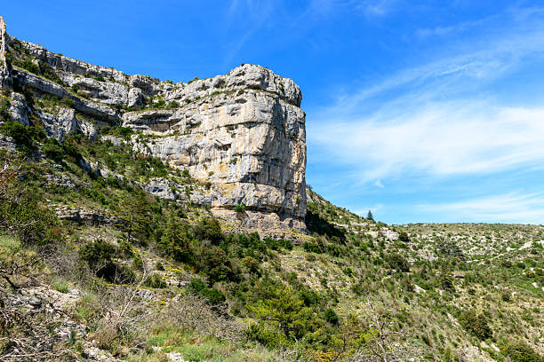 Imposing vertical limestone formation rises above a rugged, shrub-dotted hillside in the Cirque de Navacelles. Bright sunlight and a vivid blue sky enhance the sharp rock textures and wild Mediterranean vegetation.