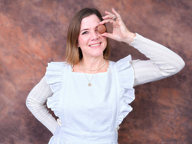 Smiling adult woman wearing an apron and holding a macaron in front of her eye, having fun