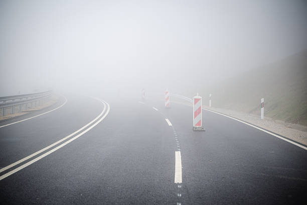 A freshly paved road winds into dense fog, with temporary red-and-white traffic barriers placed along the center line. The thick mist limits visibility and adds a moody, atmospheric feel. Road markings and guardrails emphasize caution in these challenging weather conditions.