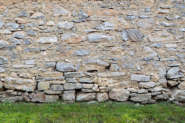 A rustic wall made of irregular stones and clay mortar, showing signs of age and weathering. At the base of the wall, fresh green grass adds contrast to the earthy textures. The image evokes a rural atmosphere and traditional construction methods.