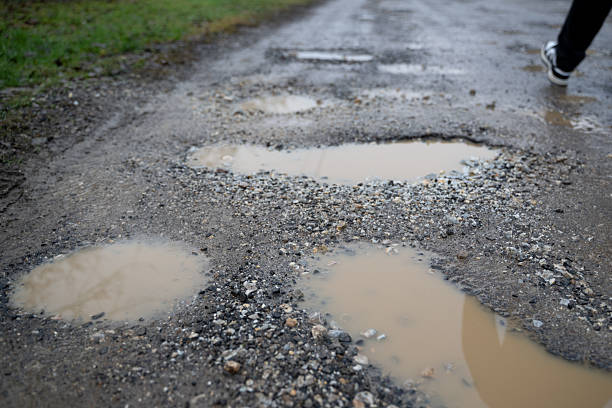 A wet gravel road is filled with multiple muddy potholes reflecting the overcast sky. In the background, a person walks away, partially visible with one foot mid-step. The image conveys a raw, rural atmosphere and highlights the effects of weather and wear on unpaved roads.
