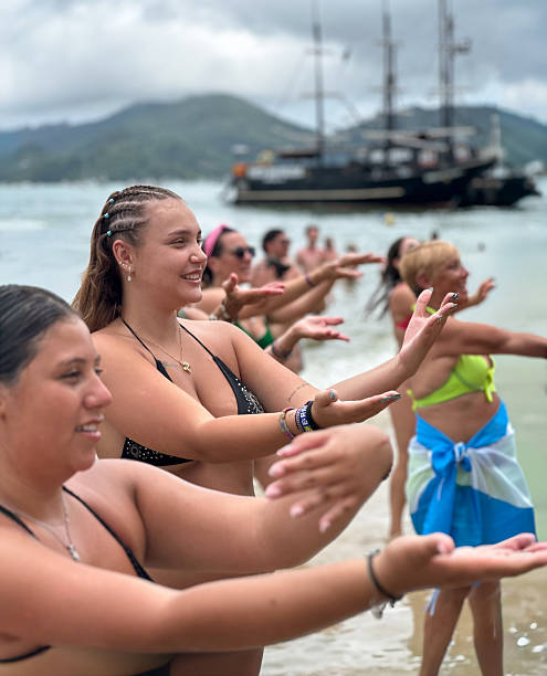 Tourists of different ages take part in a guided dance and entertainment activity organized by beach workers with a pirate tour boat anchored offshore in the background. Bombinhas, Santa Catarina, Brazil. 12/31/2025
