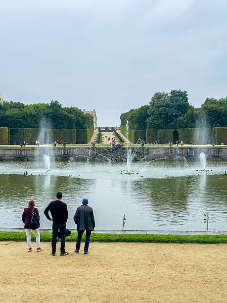 Vertical photo of three people standing on a gravel pathway looking out over a lake at sprays of water, forest groves and an avenue leading to the Palace of Versailles.