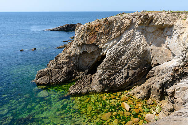 Sharp coastal rocks rise above crystal clear green-blue water at Pointe Beg-An-Aud in Saint Pierre Quiberon, with sunlight highlighting textured stone and submerged boulders.