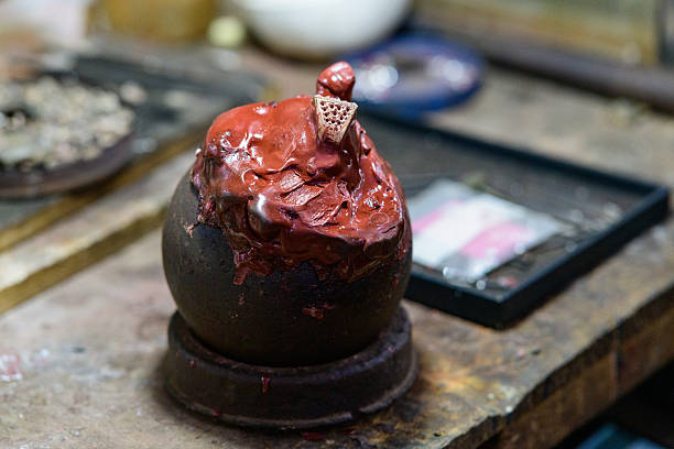 A close-up of a red wax model for jewelry making, fixed atop a dark spherical base on a worn wooden workbench in Battambang, Cambodia. The scene highlights the textured wax, intricate metalwork, and artisanal process in a traditional craft environment.