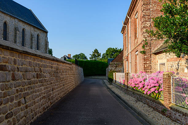 Narrow street in Saint Sylvain bordered by a textured stone wall and a red brick house with vibrant pink hydrangea flowers, bathed in warm evening sunlight under a clear blue sky.