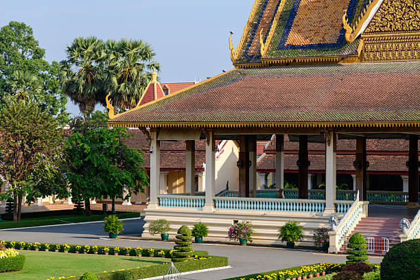 Vibrant, ornate temple-style building in Phnom Penh featuring turquoise accents, gold detailing, and monks in orange robes walking nearby. Bright sun highlights decorative elements and architectural grandeur in an urban setting.