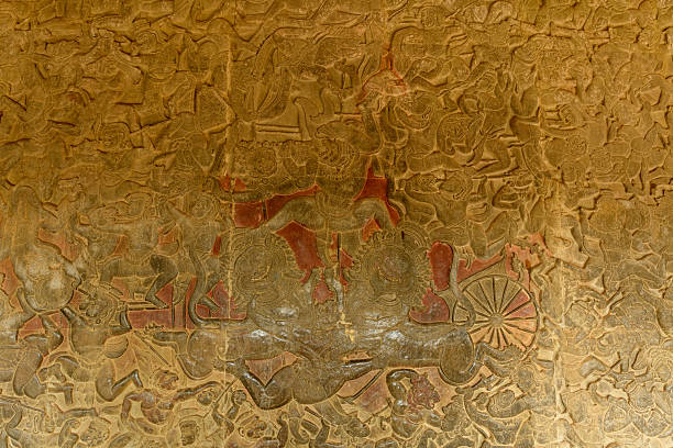 Detailed sandstone bas relief depicts a dynamic battle scene on the honor terrace of Angkor Vat. The weathered carvings display layered figures, ornate headdresses, and textured surfaces, showcasing the artistry of ancient Khmer craftsmanship.