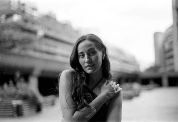 Grainy black and white analog photograph capturing a young woman with long hair, posing thoughtfully in a softly blurred urban backdrop, gazing directly at the camera
