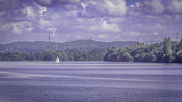 A wide view over Lake Kemnader; a single white sailboat crosses on the glittering water in front of a backdrop of dense forests and high-voltage masts under an imposing cloud sky.