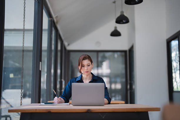 istock Young adult woman intently working on her laptop computer while also taking notes in a notebook, concentrating on her business tasks in a modern co-working office space 2255177971