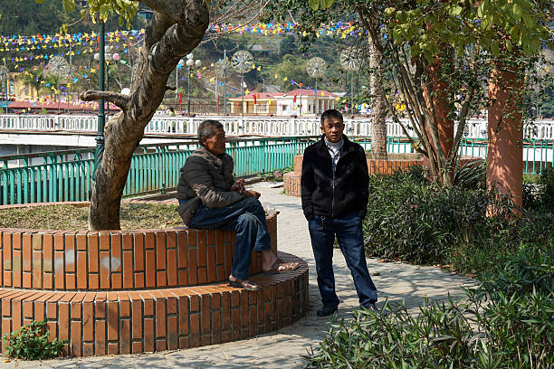 Two Asian men having a casual conversation while resting in a public park with colorful decorations and greenery in the background.