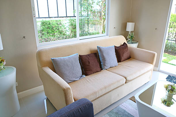 Fragment of a room with a sofa and table, a window with tulle. Living room with large sofa, pillows, coffee table, laminate flooring.