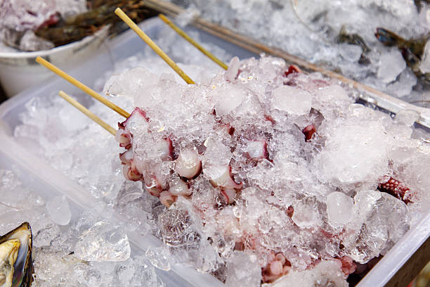 A tray of octopus is sitting in a container of ice. The octopus is covered in ice and is being served with skewers