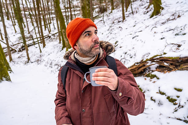 Middle-aged man in an orange hat and winter coat pauses on a snowy forest hike, holding a steaming coffee cup and gazing into the tranquil woodland for a quiet break