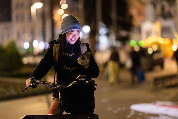 istock Smiling Girl Riding a Bicycle at Night While Checking Her Smartphone, Warm Winter Gear, Soft City Lights 2255193715