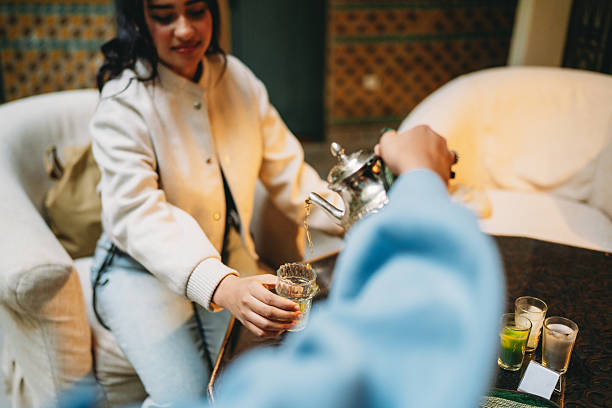 Young adult woman sitting on a couch, receiving traditional mint tea poured from a silver teapot in a cultural setting