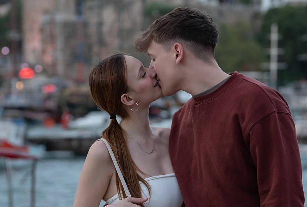 A young man and woman in a romantic embrace, sharing a kiss outdoors by a scenic harbor