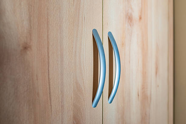 This detailed image features the front panels of a light-colored wooden cabinet or wardrobe. The focus centers on two sleek, arc-shaped metal handles mounted vertically where the double doors meet. The surface of the doors displays a smooth, light brown wood grain pattern—resembling beech or maple—which contrasts warmly with the cool, satin silver finish of the hardware. The clean lines and simple geometry suggest a contemporary or minimalist furniture design.