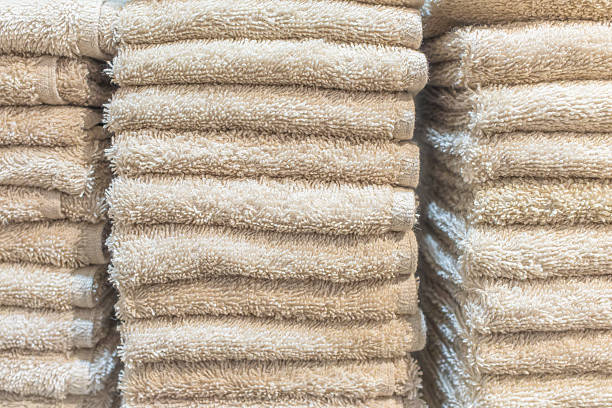 This close-up image displays neat vertical stacks of folded bath towels arranged side-by-side. The towels appear to be a distinct gray color, though they take on a warmer, brownish hue due to the ambient yellow lighting. The close perspective emphasizes the detailed, looped texture of the terry cloth fabric, while the repetitive horizontal lines of the folds create a sense of order and soft comfort.