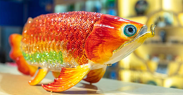 This close-up captures a beautifully crafted Arowana (Dragon Fish) figurine, likely made of Liuli (traditional Chinese crystal glass). The sculpture features a vibrant interplay of amber, red, and green hues that flow and blend seamlessly across the body—a hallmark of the Liuli casting technique. The surface is textured with a glistening, scale-like finish, while gold-toned details on the fins, barbels, and mouth accentuate its regal appearance, symbolizing wealth and prosperity.