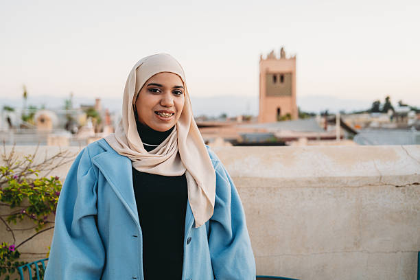 Relaxed young woman traveler wearing a hijab and light blue coat, standing on a rooftop in Marrakech city