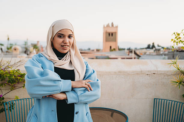 Young North African woman with braces wearing hijab and blue coat, standing on a rooftop in a Caucasion city
