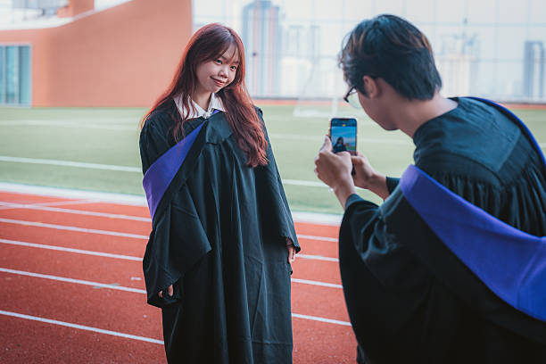 A young man is holding up a smartphone to take a picture of his friend before the graduation ceremony
