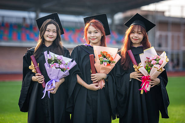 A group of three young Asian female graduates celebrate their graduation, smiling while wearing gowns and caps during a graduation ceremony at a sports field.