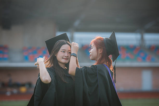 A joyful Asian female student adjusts the mortarboard of her female classmate before the graduation ceremony