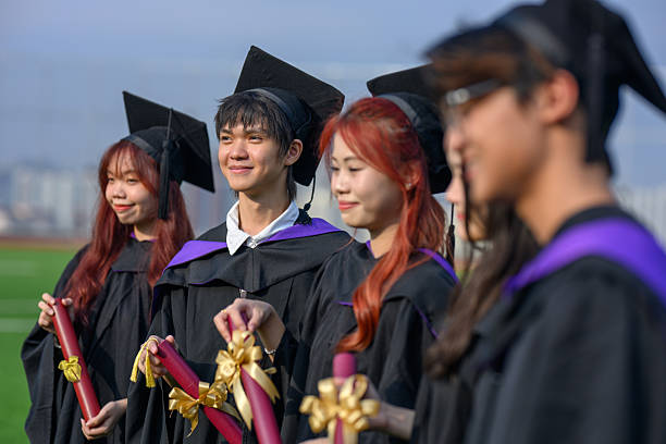 A celebration of graduation, education, and success: a young female student graduates from university outdoors. She stands with her graduating group, holding her diploma and certificate after completing her studies.