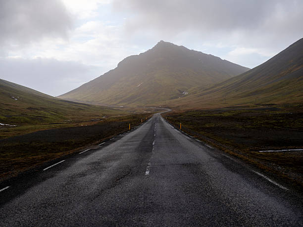 A long, straight asphalt road stretches through a vast, desolate landscape toward rugged mountains under a dramatic and cloudy sky in rural Iceland.