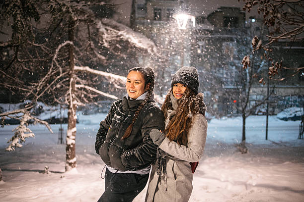 Two young women standing outdoors together at night during snowfall. Wearing winter coats and hats, smiling while snow falls around them in an urban setting. City life