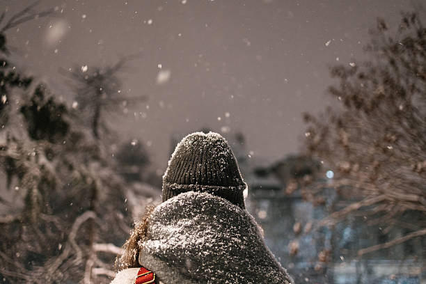 Rear view of a woman wearing winter hat and coat standing outdoors at night during snowfall. Snow-covered clothing, urban winter scene, cold weather concept.