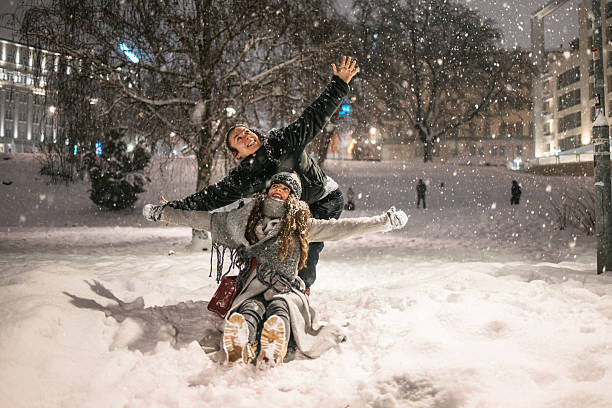 Two young women playing together outdoors in the city at night during snowfall. Sitting and kneeling in the snow with arms outstretched, wearing winter clothing, urban winter scene.