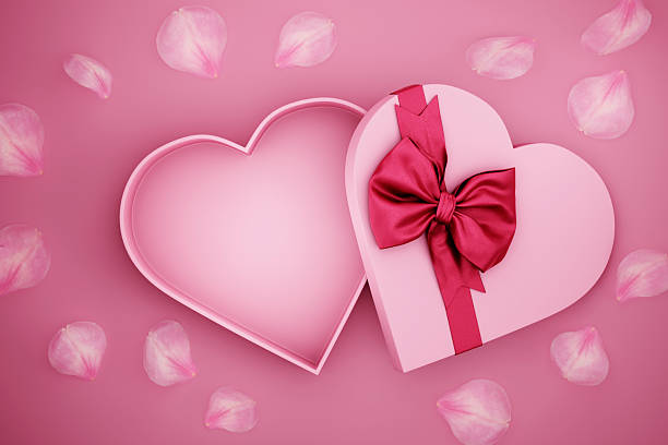 Open heart shaped present box wrapped with red bow tie and rose flower petals on pink background. Valentine's day giftbox for luxury product mockup. Mother's day, woman, birthday, anniversary holiday celebration.
