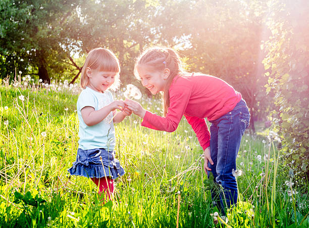 Two Little Girls Blowing Dandelions In A Spring Meadow