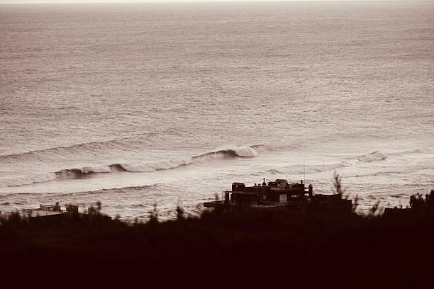 A dramatic landscape shot of the restless ocean during a cold, overcast day. The image captures powerful waves with white foam breaking near the shore, seen from a distance with a dark, moody atmosphere. This scene perfectly illustrates the raw power of nature, solitude, and the melancholic beauty of the sea in the off-season.