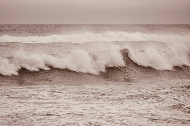 A dramatic and atmospheric shot of the ocean during a stormy winter day. The image captures a large, powerful wave with white foam as it breaks, set against a dark and moody sky. The muted tones and the spray from the water evoke a sense of the raw power and solitude of the sea. Perfect for cinematic backgrounds, environmental themes, or editorial content related to nature's strength.