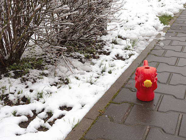 A playful red elephant watering can rests on a snowy sidewalk, symbolizing the transition to spring. Perfect for seasonal gardening, outdoor living, and unpredictable weather themes.