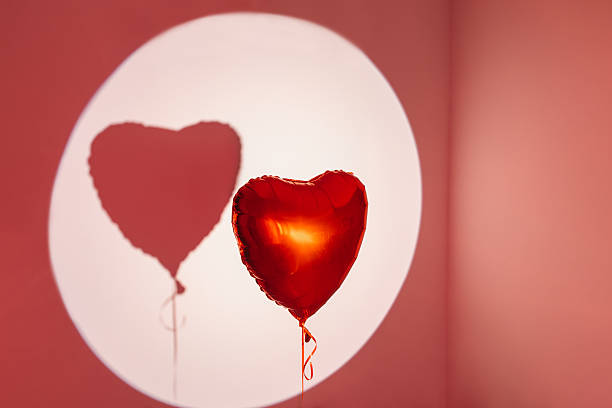Red heart-shaped balloon floating against a red background with visible shadow. Minimalistic love and romance concept with studio lighting.