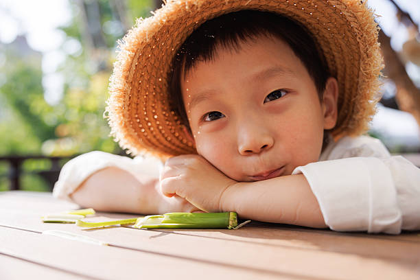 This is an Asian boy wearing a straw hat, around 5 years old. Playing outdoors in summer, he felt a bit awkward and rested on the table