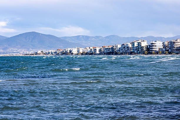 Wind-driven waves roll across the Thermaic Gulf toward the coastal town of Peraia, with dense seaside apartment buildings lining the shore and low mountains rising in the background.