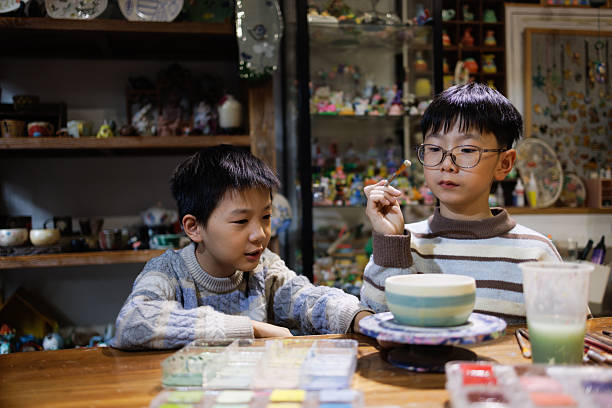 These are two little Asian Boys, about 10 years old. They are brothers, dressed in casual clothes, and are coloring their pottery bowls in a handicraft workshop. They are very happy and serious.
