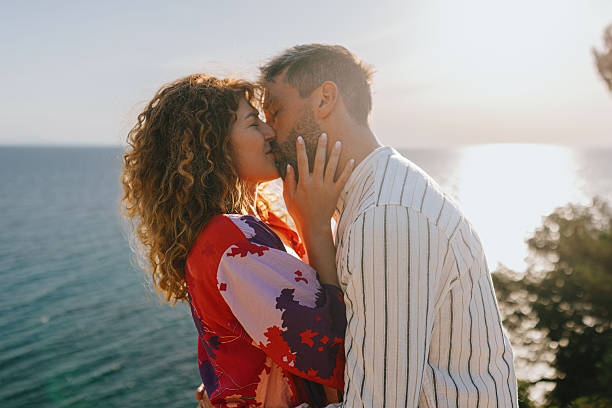 Silhouette of man kissing and hugging woman at sunset against background of moon in sky. Female kisses male and enjoy evening on summer day. Couple in love on seashore in sand sea. Place for text