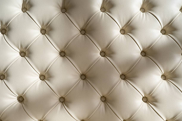Luxury tufted leather upholstery texture in soft beige tones. Close-up of a classic button-tufted sofa or headboard with diamond pattern, smooth leather surface, and elegant padding. Carriage coupler technique .