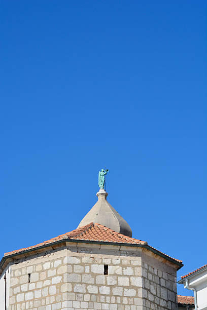 A copper statue of an angel blowing a trumpet stands atop the stone tower of the Cathedral of the Assumption of the Blessed Virgin Mary in Krk under a clear blue sky.