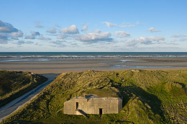 A solitary concrete blockhouse sits nestled among windswept green dunes overlooking the wide sandy shore of Utah Beach, with gentle waves and scattered clouds under a clear blue sky. The scene combines rugged textures with a peaceful, open coastal atmosphere.