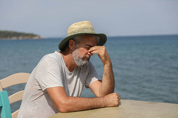 Middle Aged Man Wearing Straw Hat Sitting By The Sea With Hand Covering Eyes, Expressing Stress, Exhaustion, Headache, Or Emotional Overload During Summer Vacation, Highlighting Burnout, Mental Fatigue, And Inner Tension In Relaxing Coastal Environment.