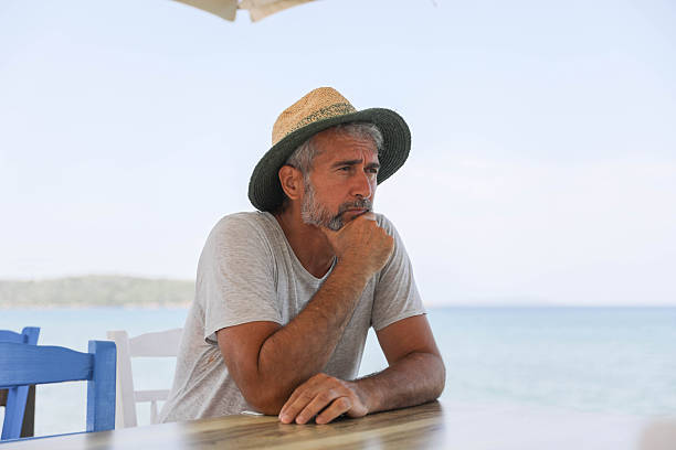 Middle Aged Man Wearing Straw Hat Sitting At Seaside Table With Hand On Chin, Showing Thoughtful Expression, Contemplation, And Quiet Reflection During Summer Day By The Sea, Highlighting Lifestyle, Mindfulness, And Emotional Balance In Coastal Environment.